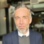 A man with grey hair and beard, wearing a blue suit jacket over a grey sweater and white dress shirt, stands indoors with a confident expression. The background is blurred but suggests a modern urban setting.