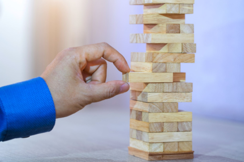 A close-up of a hand in a blue shirt carefully pulling a wooden block from a Jenga tower.