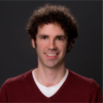 A man with curly dark hair smiles at the camera. He is wearing a dark red sweater over a white shirt. The background is dark gray.