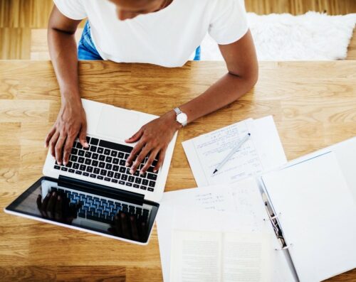 Overhead view of a person typing on a laptop on a wooden desk. Papers, a pen, and an open binder with notes about the proposal are spread around the workspace.