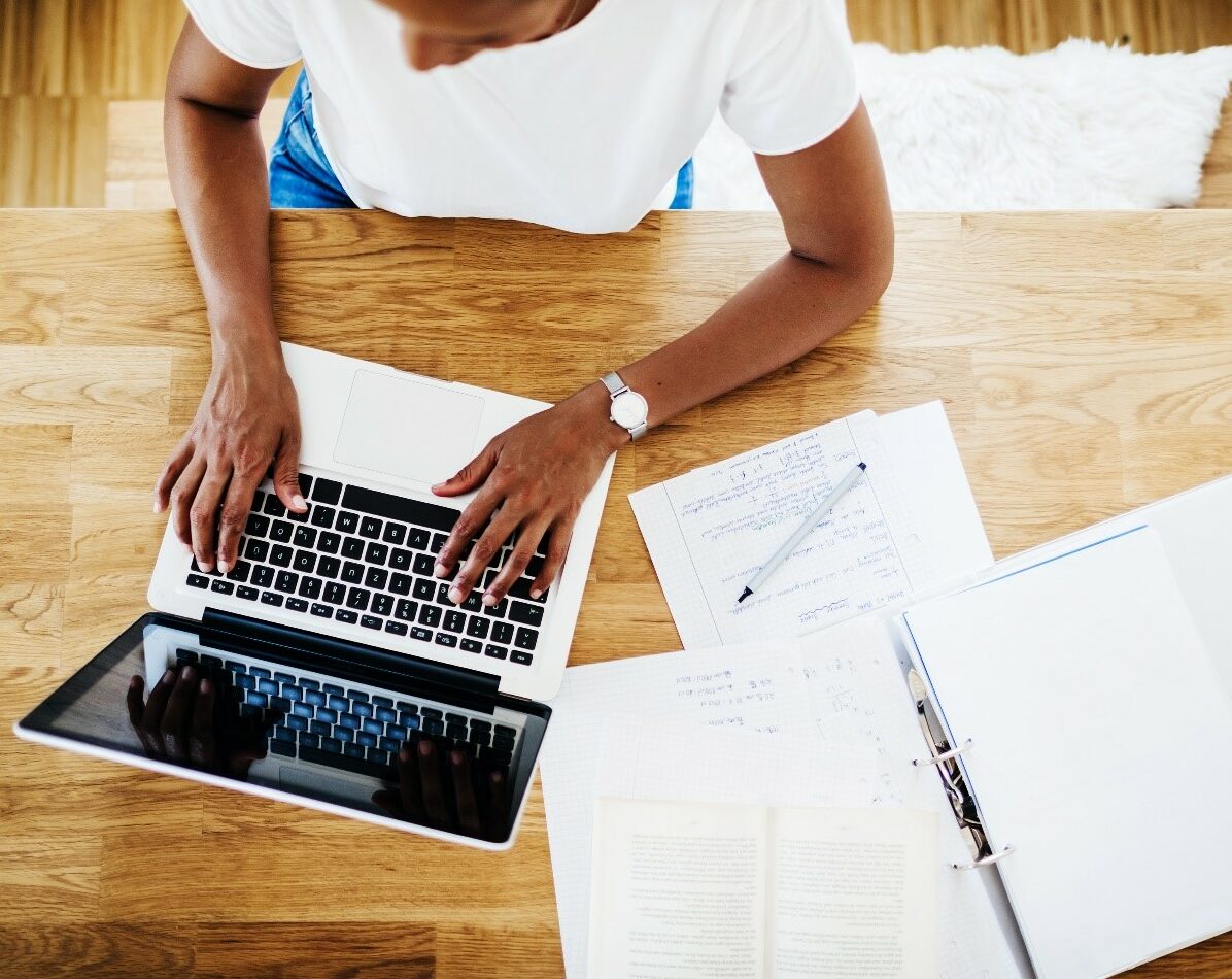 Overhead view of a person typing on a laptop on a wooden desk. Papers, a pen, and an open binder with notes about the proposal are spread around the workspace.