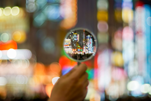 A hand holds a magnifying glass that focuses on a busy, brightly lit urban street filled with neon signs and colorful lights. The background outside the magnified area is blurred, highlighting the cityscape viewed through the magnifying glass.