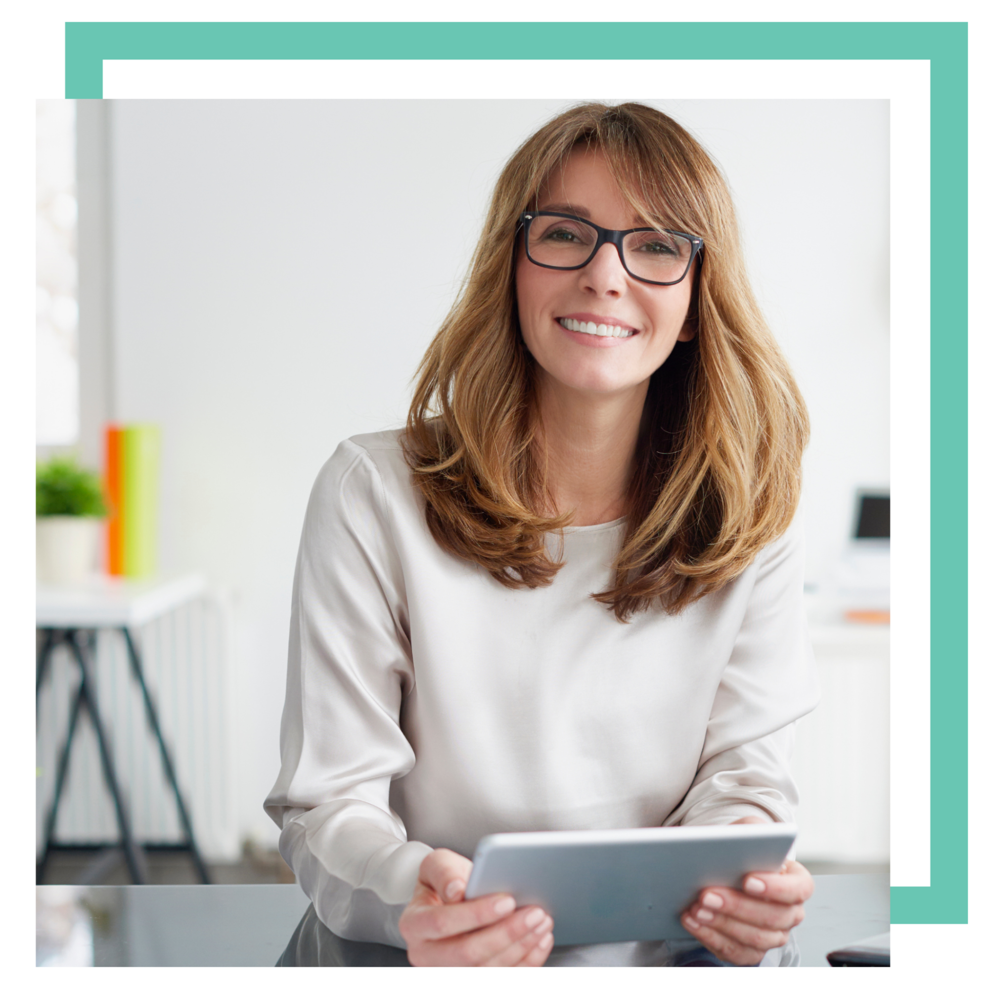 A smiling woman with long hair and glasses sits behind a desk, holding a tablet. She is wearing a light-colored blouse. The background features a bright, modern office setting with a blurred image of furniture and decor.