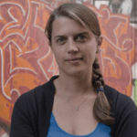 A woman with long hair, tied in a braid over her shoulder, stands in front of a colorful graffiti wall. She is wearing a blue top and a black cardigan, and looks directly at the camera with a neutral expression.