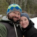 A smiling man with a beard and a colorful beanie and a woman with a headband stand close together in a snowy forest. Both are dressed in warm clothing suitable for winter weather. Trees without leaves can be seen in the background.