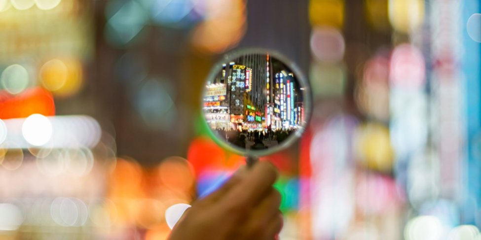 A hand holds a magnifying glass that focuses on a busy, brightly lit urban street filled with neon signs and colorful lights. The background outside the magnified area is blurred, highlighting the cityscape viewed through the magnifying glass.