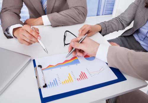 Several people in business attire sit at a desk analyzing a colorful line graph and bar chart on a clipboard, preparing for their upcoming proposal.