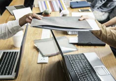 A group of people sit around a table with laptops, notebooks, and documents. Two people in the foreground are passing a proposal folder. Various papers and office supplies are scattered on the table, and a small potted plant is visible in the center.