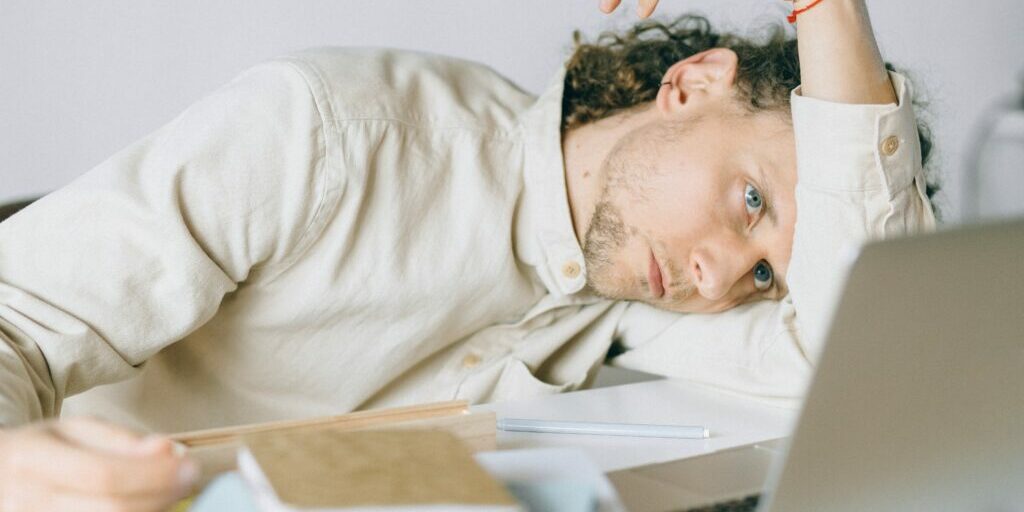 A person with long curly hair and a neutral expression is resting their head on their arm at a desk. They are wearing a light-colored shirt and are surrounded by notebooks, a pen, and a laptop. The scene suggests feeling tired or overwhelmed.