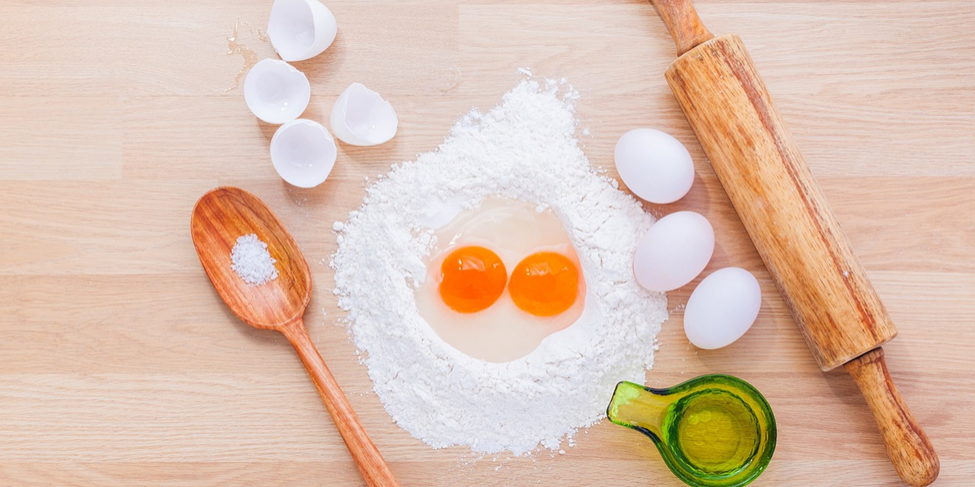 A kitchen counter with flour, two cracked eggs, a wooden spoon with salt, a green glass cup with oil, a rolling pin, and three whole eggs. Eggshells are scattered nearby. The ingredients are arranged for baking.