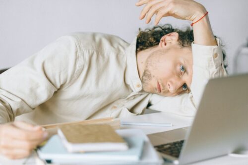 A person with long curly hair and a neutral expression is resting their head on their arm at a desk. They are wearing a light-colored shirt and are surrounded by notebooks, a pen, and a laptop. The scene suggests feeling tired or overwhelmed.