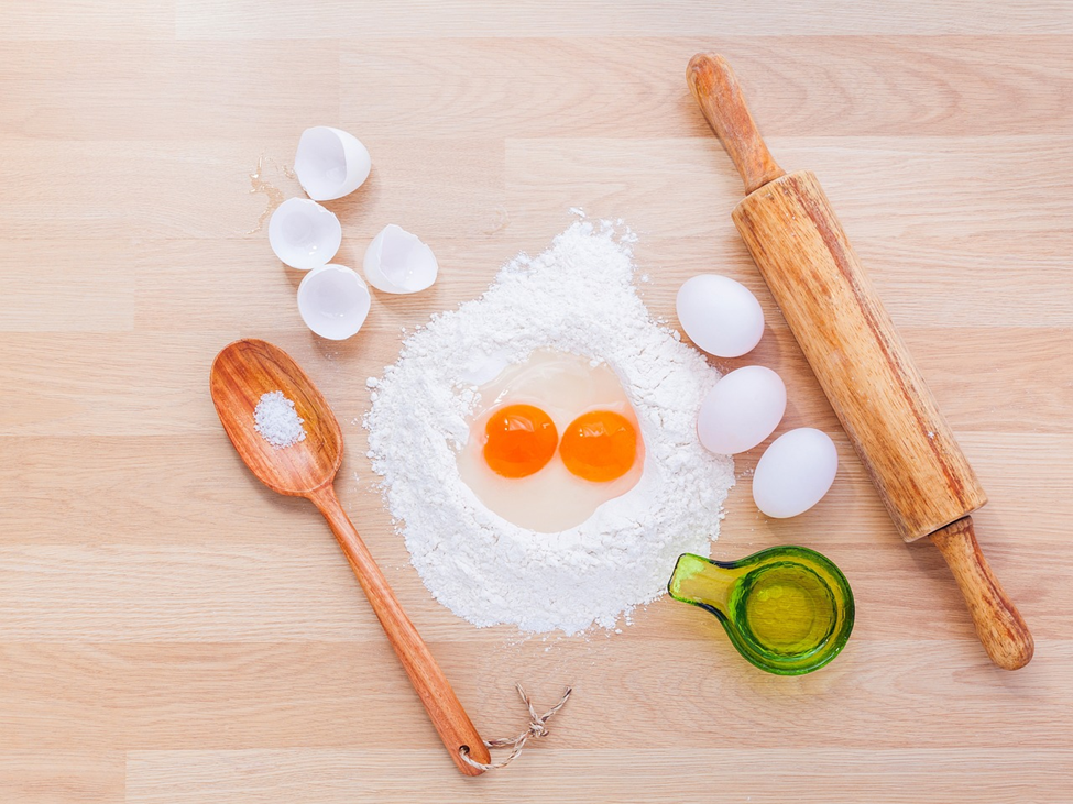 A kitchen counter with flour, two cracked eggs, a wooden spoon with salt, a green glass cup with oil, a rolling pin, and three whole eggs. Eggshells are scattered nearby. The ingredients are arranged for baking.